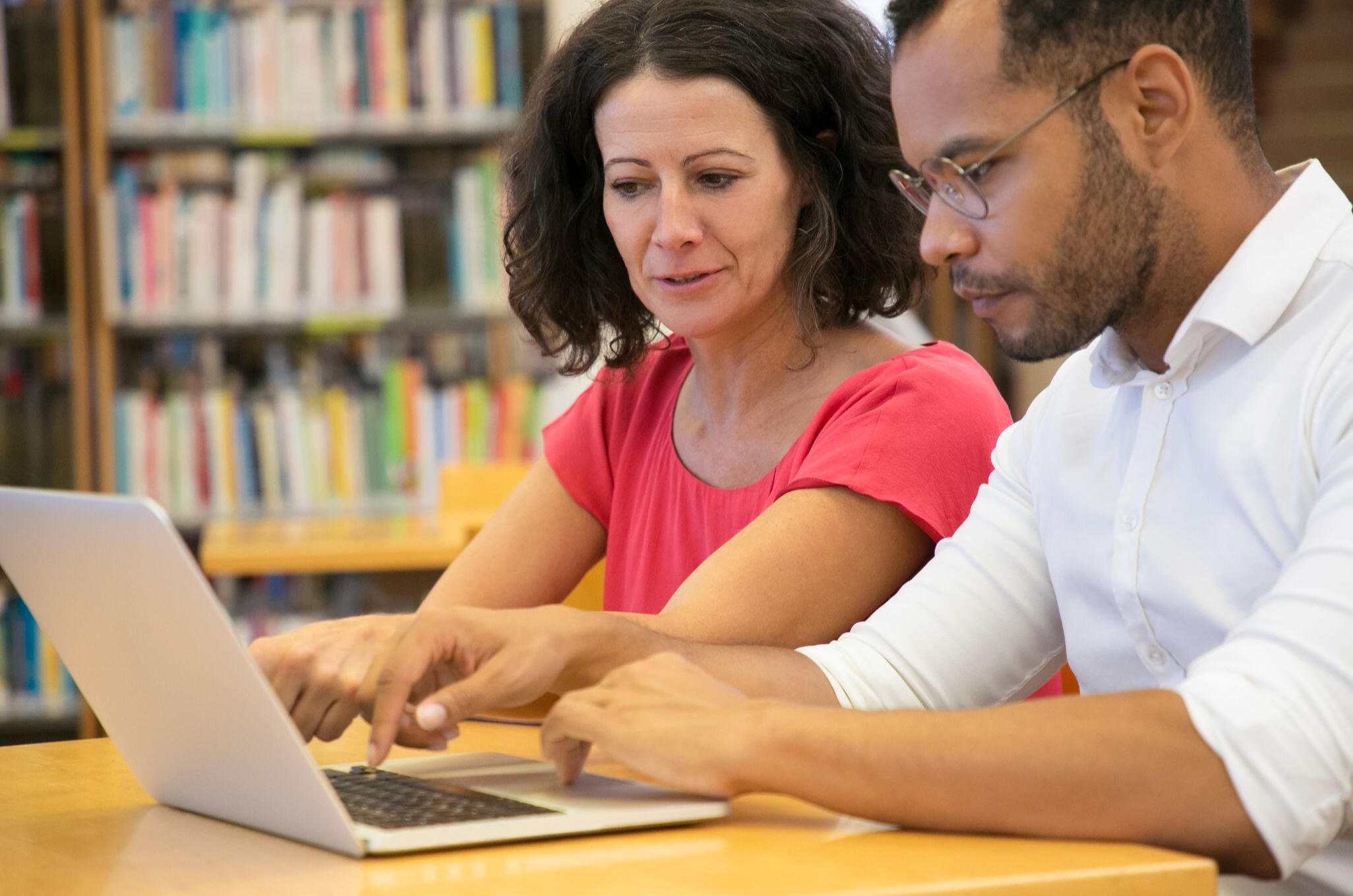 Picture of two adults, one a white woman with dark hair, the other a black man with glasses and a short beard. They are at a desk interacting with a laptop computer.